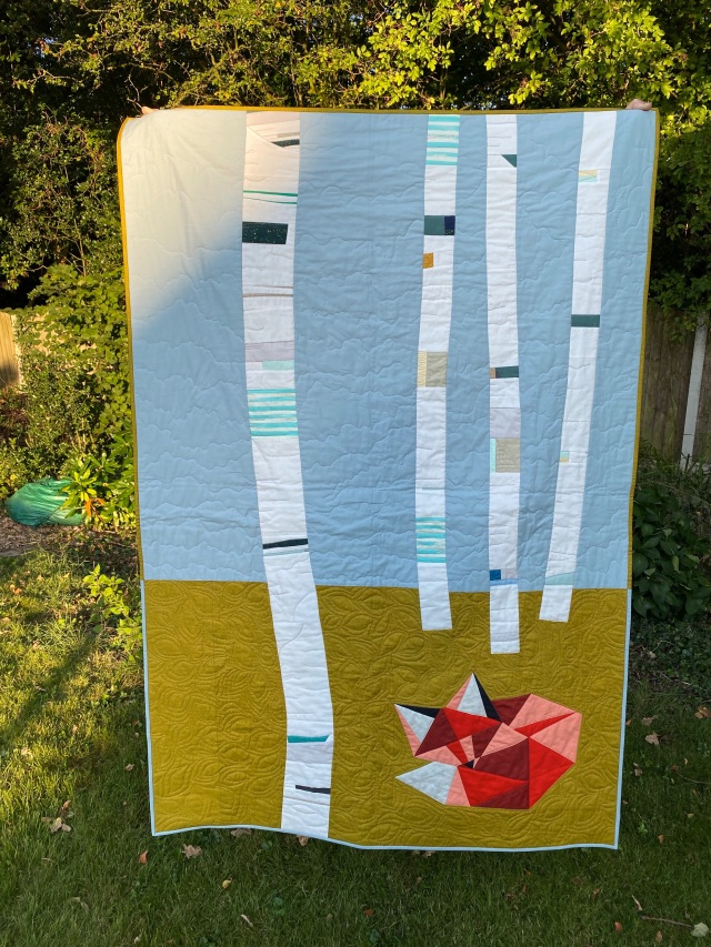 A modern pieced quilt in held up in a garden. The quilt depicts a little fox curled up on a green background, with four birch trees against a blue sky.